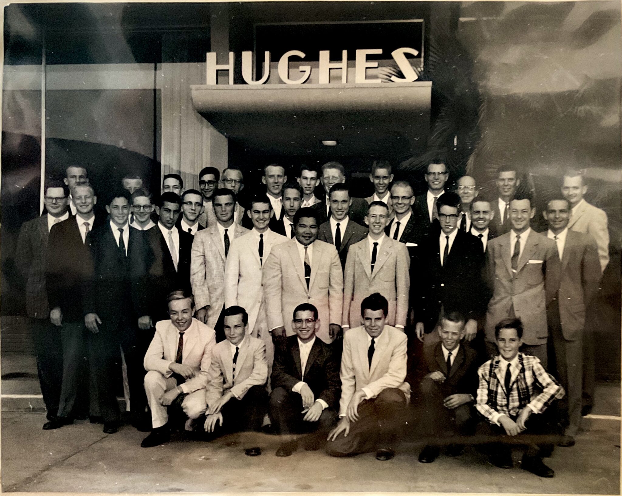 A group of men in suits and ties stand and kneel in rows, embodying a legacy of mission and history, in front of a building adorned with a