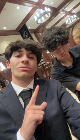Two young men indoors, one wearing a suit and pointing upwards, the other with curly hair looking down. They are in a room with wooden beams and pendant lights.